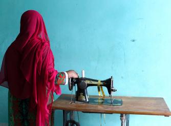 Rahima, a garment factory worker in Bangladesh, next to a sewing machine and with her back to camera